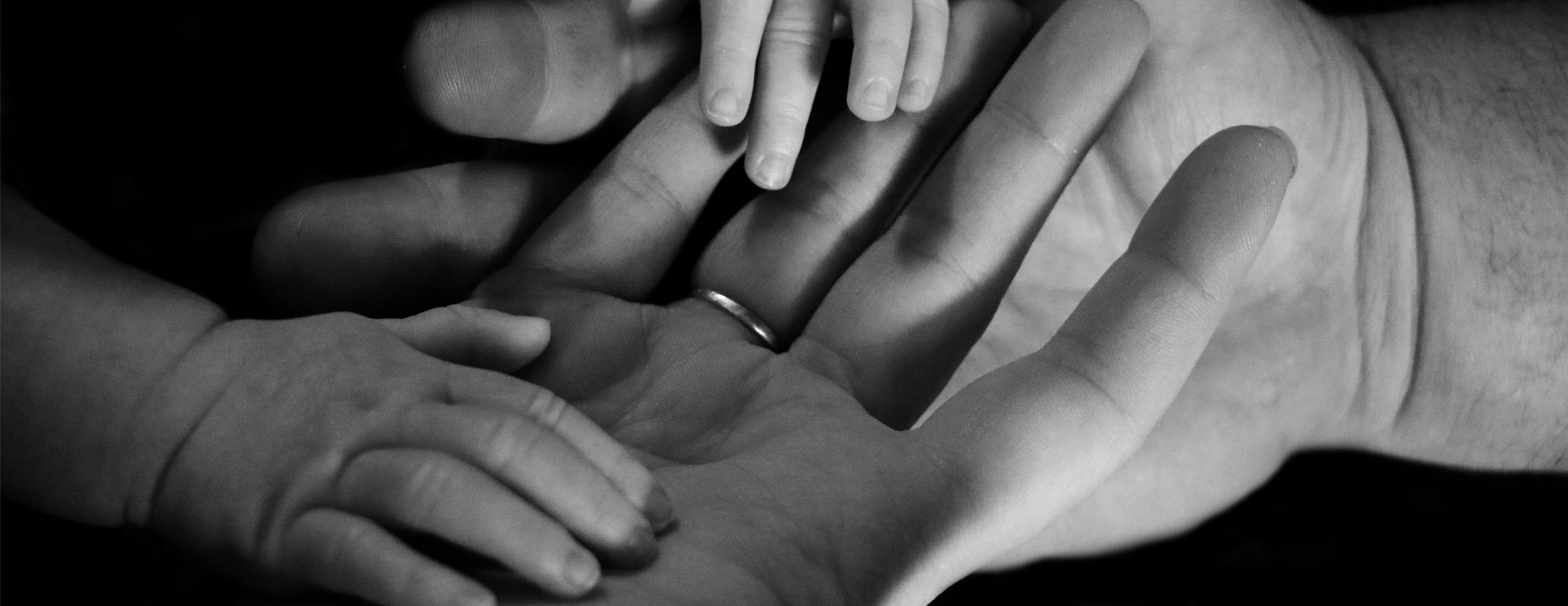 Black and white close up of adult hands holding baby hands. The adult hand has a wedding ring.