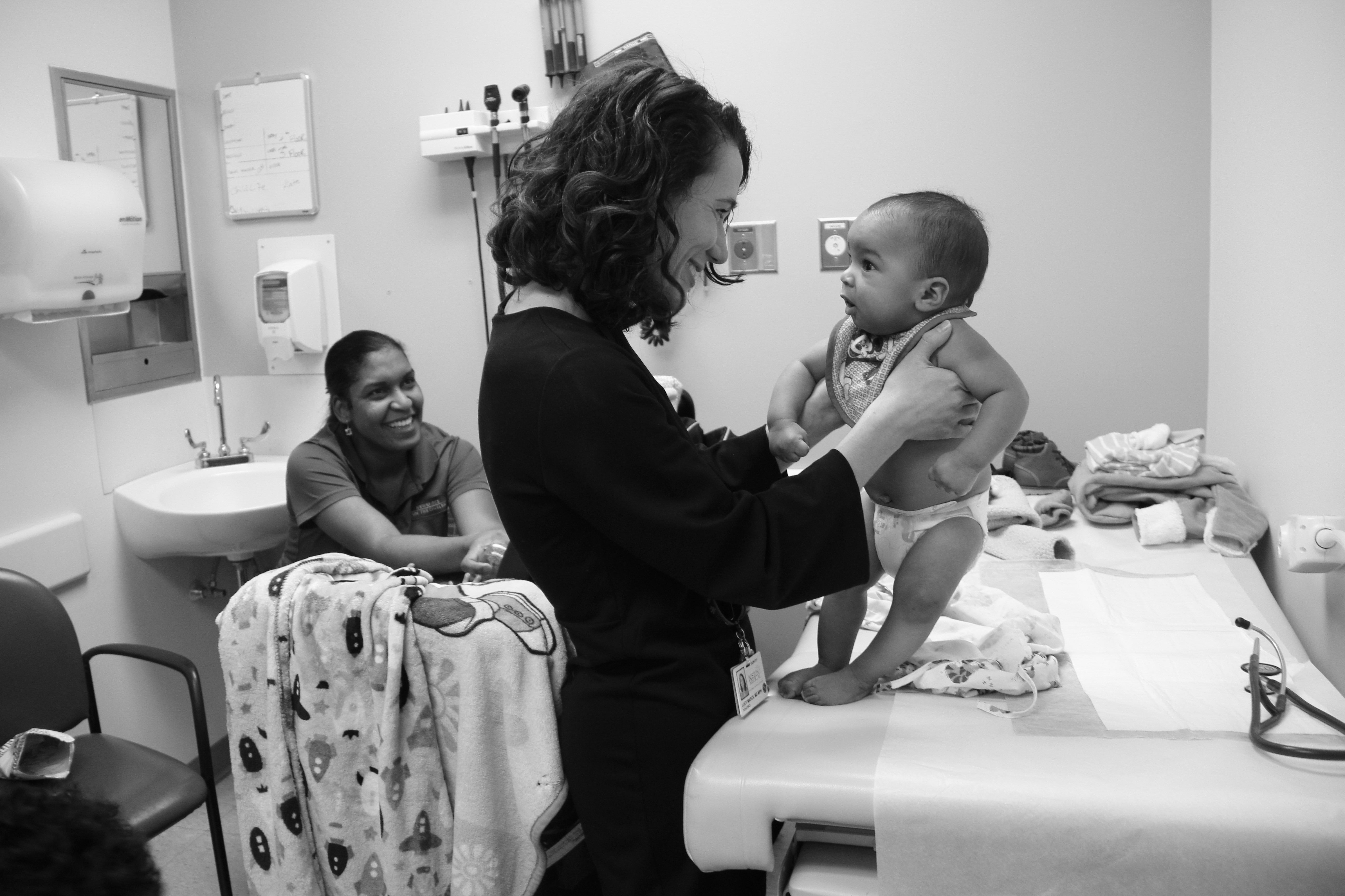 Lucy Marcil holding a young baby in a BMC clinic while mom watches with a smile from the background
